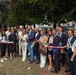A formal ribbon-cutting ceremony is taking place outdoors in what appears to be an Italian municipality, with officials and dignitaries holding a ribbon in the Italian national colors (green, white, and red). Several participants are wearing ceremonial sashes also in Italian tricolor colors, indicating they are likely elected officials or mayors. A municipal banner/gonfalon is visible on the left side of the image.