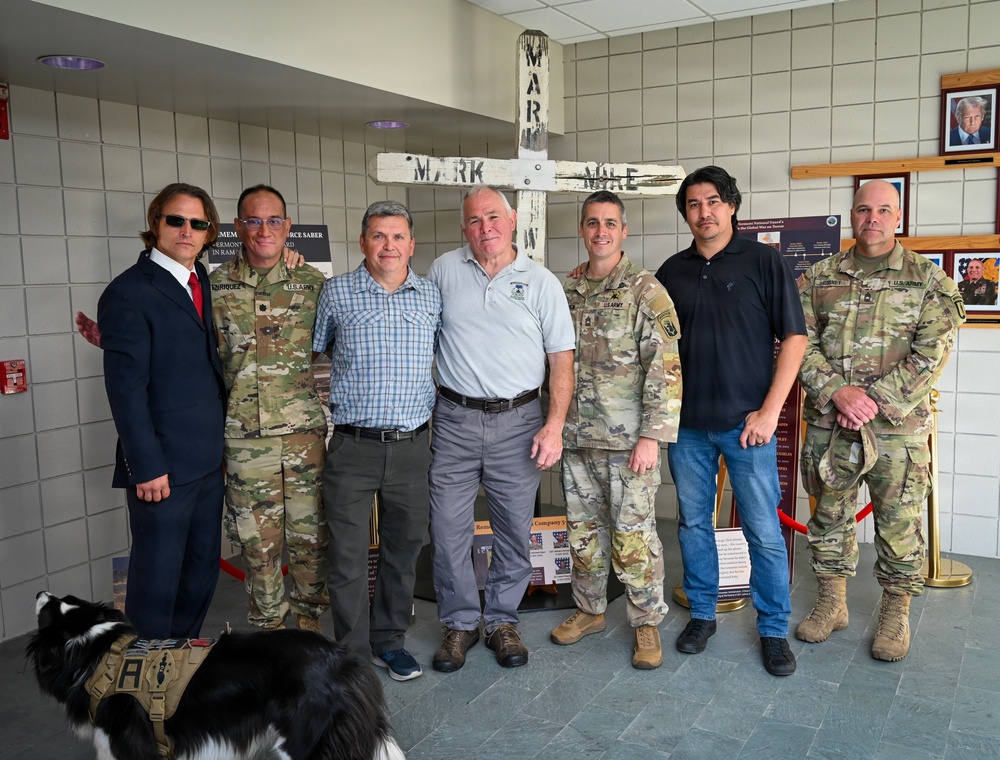 A group of seven individuals pose for a photo inside what appears to be a military facility or memorial hall. Four of the individuals are wearing U.S. Army camouflage uniforms, while three are in civilian attire. They are standing in front of a white wooden cross memorial bearing the name 'MARK' and 'MIKE', with military display boards and a framed portrait visible in the background.