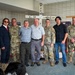 A group of seven individuals pose for a photo inside what appears to be a military facility or memorial hall. Four of the individuals are wearing U.S. Army camouflage uniforms, while three are in civilian attire. They are standing in front of a white wooden cross memorial bearing the name 'MARK' and 'MIKE', with military display boards and a framed portrait visible in the background.