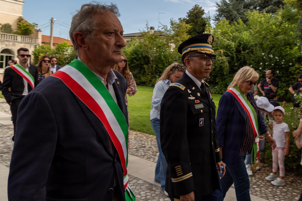 A formal outdoor procession featuring Italian civic officials wearing the traditional tricolor sashes (fascia tricolore) alongside a U.S. Army officer in dress uniform. The setting appears to be a garden or park adjacent to a historic building, suggesting a commemorative or diplomatic ceremony. The gathering includes civilians and onlookers in the background.