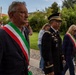 A formal outdoor procession featuring Italian civic officials wearing the traditional tricolor sashes (fascia tricolore) alongside a U.S. Army officer in dress uniform. The setting appears to be a garden or park adjacent to a historic building, suggesting a commemorative or diplomatic ceremony. The gathering includes civilians and onlookers in the background.