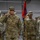 A group of U.S. Army soldiers are standing in formation inside what appears to be a military tent or temporary facility. The lead soldier, identified by his name tape as 'ROGERS,' is rendering a salute while wearing captain rank insignia. A guidon bearer stands behind him holding a red and blue unit flag.