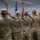 A group of U.S. Army soldiers in OCP (Operational Camouflage Pattern) uniforms are standing at salute inside a large tent or temporary structure. The soldiers are facing toward flags, including the American flag and what appears to be a state flag, suggesting a formal military ceremony. Name tapes visible on the caps include 'ROGERS', 'KING', and 'KAINE'.