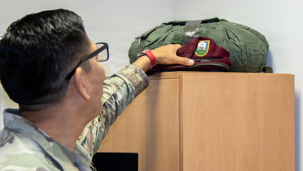 A soldier in camouflage uniform is placing or retrieving a maroon beret on top of a wooden cabinet or filing unit. The beret has a visible flash/patch on it, and an olive drab military bag is also resting on top of the cabinet. The setting appears to be an indoor military office or administrative space.