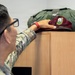 A soldier in camouflage uniform is placing or retrieving a maroon beret on top of a wooden cabinet or filing unit. The beret has a visible flash/patch on it, and an olive drab military bag is also resting on top of the cabinet. The setting appears to be an indoor military office or administrative space.