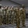 A group of U.S. Army soldiers in OCP (Operational Camouflage Pattern) uniforms are standing at attention and rendering a salute during a formal military ceremony held inside a large tent or temporary structure. One soldier in the foreground is holding a unit guidon flag with blue and red panels. The ceremony appears to be a formal formation event such as a change of command, activation, or awards ceremony.