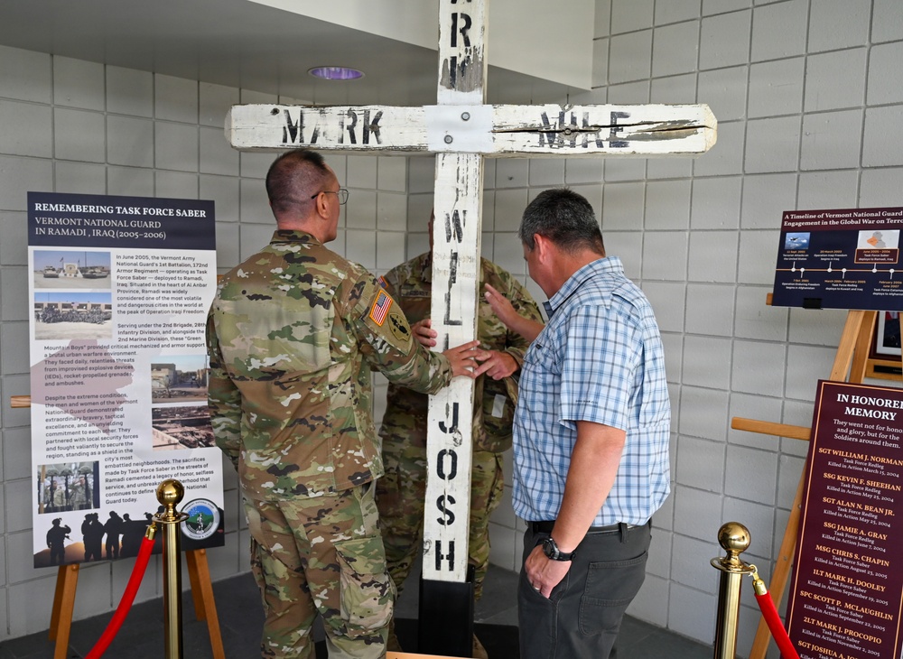 Two individuals, one in military camouflage uniform and one in civilian clothing, are examining or handling a large white wooden cross with names painted on it. The setting appears to be a memorial display, with informational boards visible in the background referencing 'Task Force Saber' and the Vermont National Guard in Ramadi, Iraq (2005-2006). The cross bears names including 'MARK,' 'MIKE,' and 'JOSH,' suggesting it is a memorial artifact honoring fallen service members.