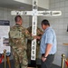 Two individuals, one in military camouflage uniform and one in civilian clothing, are examining or handling a large white wooden cross with names painted on it. The setting appears to be a memorial display, with informational boards visible in the background referencing 'Task Force Saber' and the Vermont National Guard in Ramadi, Iraq (2005-2006). The cross bears names including 'MARK,' 'MIKE,' and 'JOSH,' suggesting it is a memorial artifact honoring fallen service members.