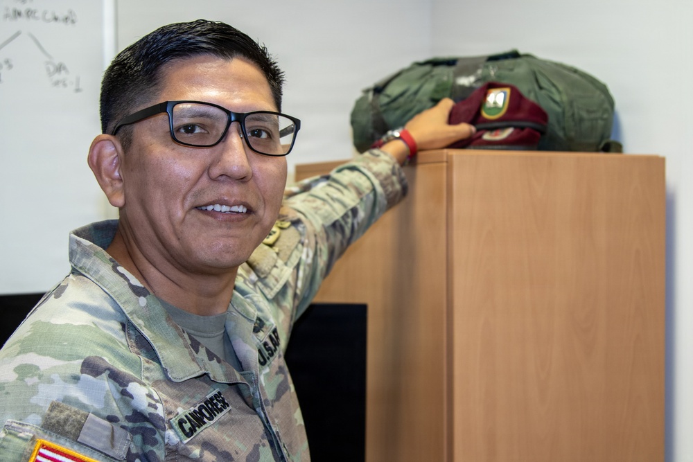 A U.S. Army soldier in OCP (Operational Camouflage Pattern) uniform is posing next to a wooden cabinet or shelf, resting his hand on top of it. On top of the cabinet sits a maroon beret with a visible flash/crest and what appears to be a green flight or kit bag. The soldier is wearing glasses and has a name tape visible on his uniform.