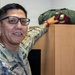 A U.S. Army soldier in OCP (Operational Camouflage Pattern) uniform is posing next to a wooden cabinet or shelf, resting his hand on top of it. On top of the cabinet sits a maroon beret with a visible flash/crest and what appears to be a green flight or kit bag. The soldier is wearing glasses and has a name tape visible on his uniform.