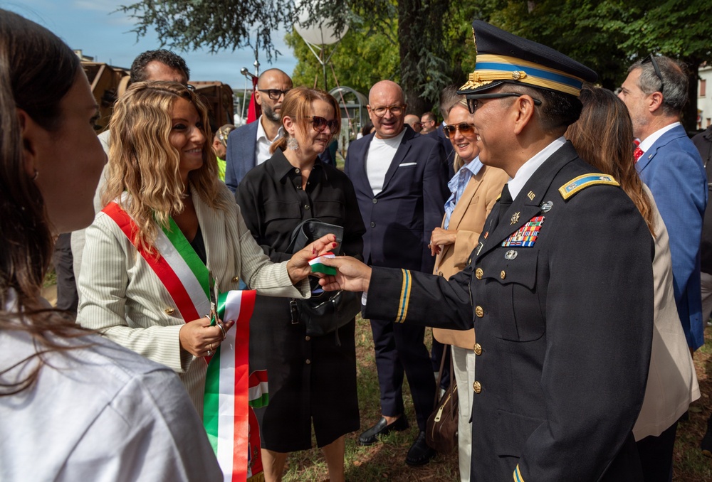 A formal outdoor ceremony or reception showing a U.S. Army officer in dress uniform greeting a woman wearing an Italian tricolor sash, indicating she holds an official Italian civic role such as a mayor or municipal official. The gathering includes multiple attendees in business attire, suggesting a diplomatic or commemorative event. The setting appears to be a park or outdoor public space in Italy.