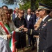 A formal outdoor ceremony or reception showing a U.S. Army officer in dress uniform greeting a woman wearing an Italian tricolor sash, indicating she holds an official Italian civic role such as a mayor or municipal official. The gathering includes multiple attendees in business attire, suggesting a diplomatic or commemorative event. The setting appears to be a park or outdoor public space in Italy.