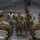 A military formation ceremony is taking place inside a large tent or temporary structure. Soldiers in OCP (Operational Camouflage Pattern) uniforms are standing at attention while two senior personnel observe from seats in the foreground. A guidon (unit flag) with red and blue colors is being held by one of the soldiers in the formation.