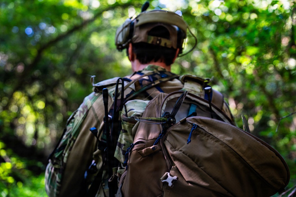 A soldier or military personnel is photographed from behind while moving through a dense, green forested environment. The individual is equipped with full tactical gear including a helmet with mounted devices, camouflage uniform, and a large tactical backpack. The image appears to depict a field operation or training exercise in a woodland setting.