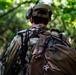 A soldier or military personnel is photographed from behind while moving through a dense, green forested environment. The individual is equipped with full tactical gear including a helmet with mounted devices, camouflage uniform, and a large tactical backpack. The image appears to depict a field operation or training exercise in a woodland setting.