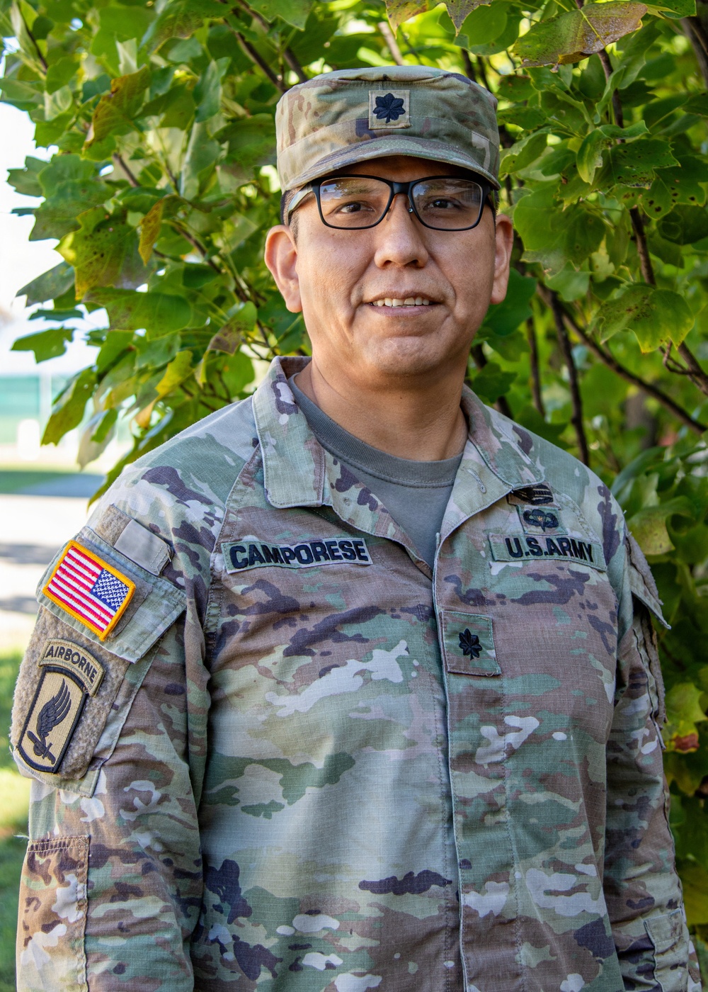 A U.S. Army Lieutenant Colonel named Camporese poses for what appears to be an official or semi-official portrait outdoors. He is wearing the Army Combat Uniform (ACU) in Operational Camouflage Pattern (OCP) with multiple visible insignia and patches. The background features green foliage consistent with a base or institutional setting.
