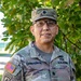 A U.S. Army Lieutenant Colonel named Camporese poses for what appears to be an official or semi-official portrait outdoors. He is wearing the Army Combat Uniform (ACU) in Operational Camouflage Pattern (OCP) with multiple visible insignia and patches. The background features green foliage consistent with a base or institutional setting.