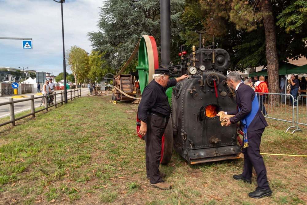 Two men are operating a vintage stationary steam engine at what appears to be an outdoor public exhibition or festival. One man dressed in black work clothes and a cap is tending to the machine, while another in a blue suit is feeding wood into the firebox, which shows visible flames. The event appears to take place along a roadside in what looks like an Italian town, evidenced by the Italian-colored flywheel and road signage.