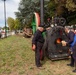 Two men are operating a vintage stationary steam engine at what appears to be an outdoor public exhibition or festival. One man dressed in black work clothes and a cap is tending to the machine, while another in a blue suit is feeding wood into the firebox, which shows visible flames. The event appears to take place along a roadside in what looks like an Italian town, evidenced by the Italian-colored flywheel and road signage.