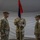Three U.S. Army soldiers are participating in a military ceremony, likely a change of command or activation ceremony, inside a tent or temporary structure. A central soldier holds a unit guidon (flag) with red and blue colors featuring a green emblem, while two soldiers flank him on either side. The soldier on the left displays general officer rank insignia, and the soldier on the right wears a '1st Infantry Division' shoulder patch.