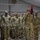 A group of U.S. Army soldiers in Operational Camouflage Pattern (OCP) uniforms are standing in formation inside a large tent or temporary structure. A soldier in the foreground is rendering a salute while holding a unit guidon (red and blue flag). The scene appears to be a formal military ceremony or formation.