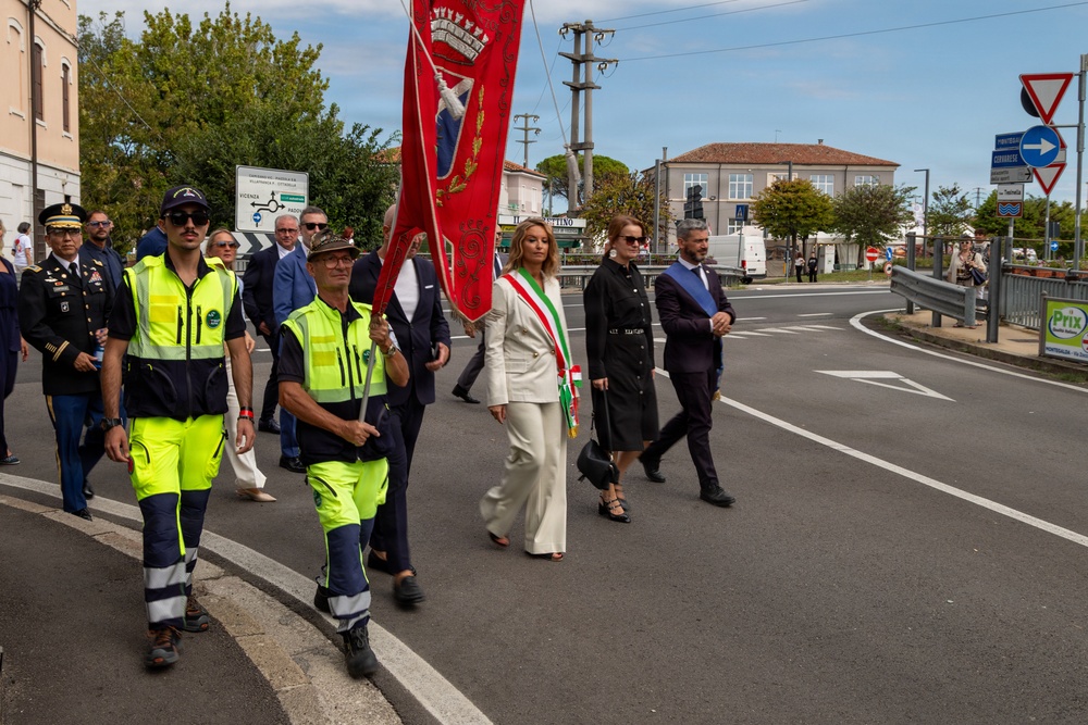 A formal civic procession is taking place on a public road in what appears to be an Italian town. The procession includes municipal police in high-visibility vests, a flag bearer carrying a red ceremonial banner, a woman wearing an Italian mayoral sash (fascia tricolore), and a US Army officer among other officials. The setting includes Italian road signs, a 'Prix' store sign, and typical northern Italian architecture.
