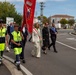 A formal civic procession is taking place on a public road in what appears to be an Italian town. The procession includes municipal police in high-visibility vests, a flag bearer carrying a red ceremonial banner, a woman wearing an Italian mayoral sash (fascia tricolore), and a US Army officer among other officials. The setting includes Italian road signs, a 'Prix' store sign, and typical northern Italian architecture.