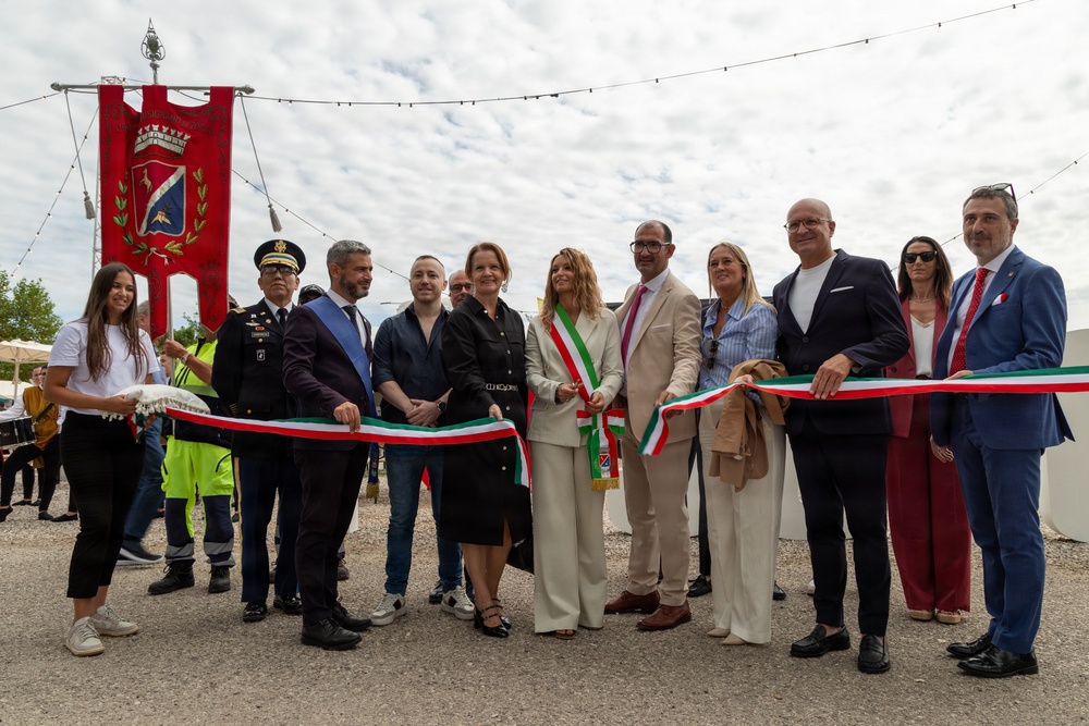 A formal ribbon-cutting ceremony is taking place outdoors, likely in Italy, as indicated by the Italian tricolor ribbon and a municipal banner (gonfalone) with a coat of arms. A group of officials and dignitaries, some wearing mayoral sashes in the Italian national colors, are gathered to inaugurate an event or facility. The setting appears to be an open-air venue with string lights overhead.