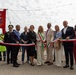 A formal ribbon-cutting ceremony is taking place outdoors, likely in Italy, as indicated by the Italian tricolor ribbon and a municipal banner (gonfalone) with a coat of arms. A group of officials and dignitaries, some wearing mayoral sashes in the Italian national colors, are gathered to inaugurate an event or facility. The setting appears to be an open-air venue with string lights overhead.