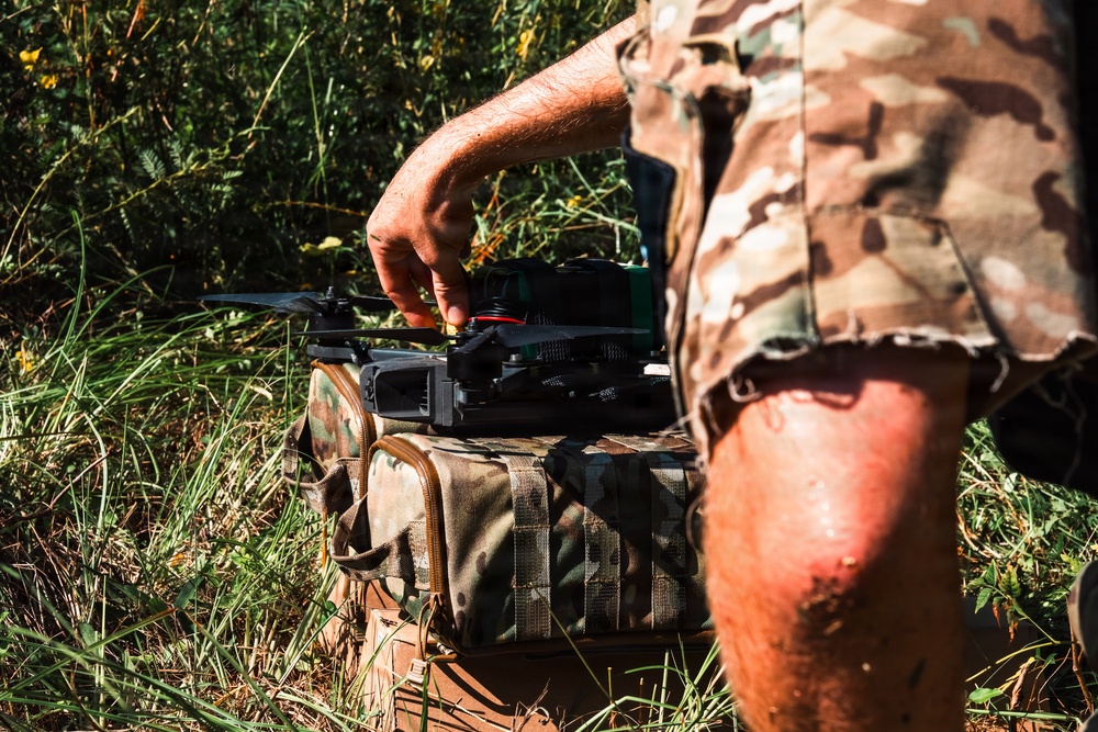 A person wearing camouflage military-style clothing is handling a small quadcopter drone placed on top of a camouflage tactical bag or case. The scene takes place outdoors in a grassy, vegetated area with natural lighting. The individual appears to be preparing or adjusting the drone for deployment.