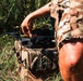 A person wearing camouflage military-style clothing is handling a small quadcopter drone placed on top of a camouflage tactical bag or case. The scene takes place outdoors in a grassy, vegetated area with natural lighting. The individual appears to be preparing or adjusting the drone for deployment.