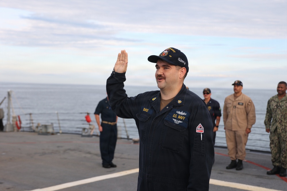 A U.S. Navy sailor in dark blue coveralls is raising his right hand, appearing to take an oath or be sworn in on the deck of a naval vessel. Several other military personnel in various uniforms are standing in the background observing the ceremony. The setting is an open ship deck with ocean and overcast sky visible in the background.