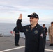 A U.S. Navy sailor in dark blue coveralls is raising his right hand, appearing to take an oath or be sworn in on the deck of a naval vessel. Several other military personnel in various uniforms are standing in the background observing the ceremony. The setting is an open ship deck with ocean and overcast sky visible in the background.