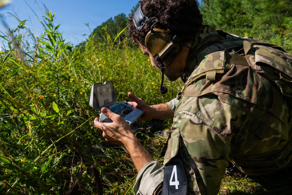 A soldier in multicam/OCP camouflage uniform is crouched in a field examining a small device or kit case. The individual is wearing tactical hearing protection and has goggles pushed up on their head, with a tactical vest/pack visible. A number '4' identifier tag is visible on their belt or lower garment, suggesting a training exercise or competition.