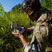 A soldier in multicam/OCP camouflage uniform is crouched in a field examining a small device or kit case. The individual is wearing tactical hearing protection and has goggles pushed up on their head, with a tactical vest/pack visible. A number '4' identifier tag is visible on their belt or lower garment, suggesting a training exercise or competition.