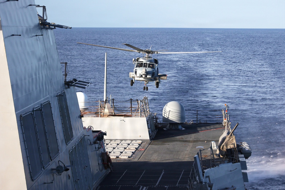 A military helicopter, appearing to be an MH-60 Seahawk, is hovering over or approaching the flight deck of a naval warship, likely a U.S. Navy destroyer or cruiser. The ship is underway at sea under clear skies with calm ocean conditions. The scene depicts a helicopter landing or departure operation on the ship's stern flight deck.