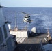 A military helicopter, appearing to be an MH-60 Seahawk, is hovering over or approaching the flight deck of a naval warship, likely a U.S. Navy destroyer or cruiser. The ship is underway at sea under clear skies with calm ocean conditions. The scene depicts a helicopter landing or departure operation on the ship's stern flight deck.
