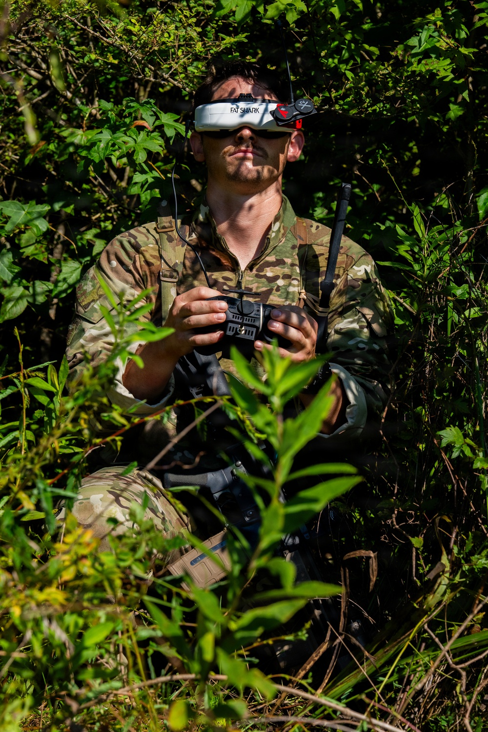 A person wearing military-style multicam camouflage clothing is positioned among dense vegetation, wearing FatShark FPV (First Person View) goggles on their forehead and holding a drone radio controller. The individual appears to be operating or preparing to operate an FPV drone, likely in a tactical or military training context. The setting is an outdoor wooded or brushy environment with bright natural sunlight.