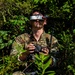 A person wearing military-style multicam camouflage clothing is positioned among dense vegetation, wearing FatShark FPV (First Person View) goggles on their forehead and holding a drone radio controller. The individual appears to be operating or preparing to operate an FPV drone, likely in a tactical or military training context. The setting is an outdoor wooded or brushy environment with bright natural sunlight.