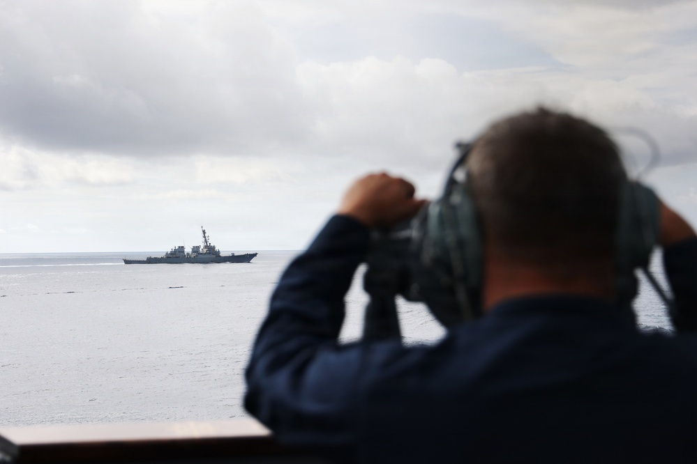 A naval personnel member is observing a distant warship through large mounted binoculars or a optical rangefinder from an elevated deck position. The distant vessel appears to be a guided-missile destroyer or similar naval combatant ship on open ocean waters. The scene suggests a maritime surveillance or monitoring operation.