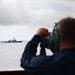 A naval personnel member is observing a distant warship through large mounted binoculars or a optical rangefinder from an elevated deck position. The distant vessel appears to be a guided-missile destroyer or similar naval combatant ship on open ocean waters. The scene suggests a maritime surveillance or monitoring operation.