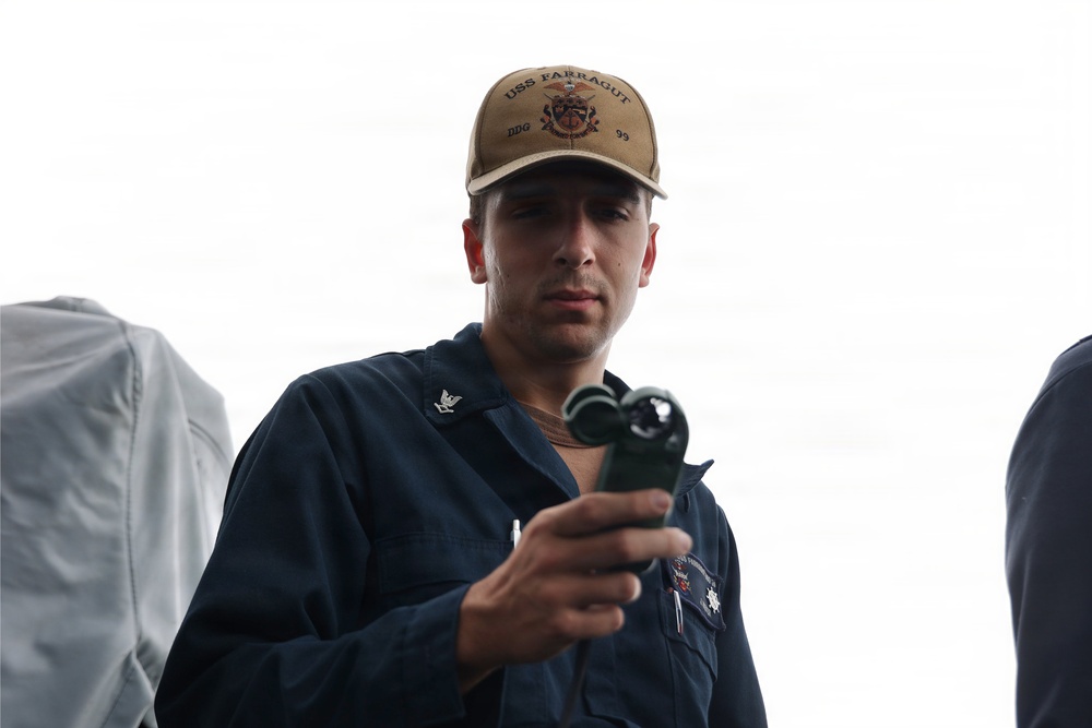 A U.S. Navy sailor wearing a USS Farragut DDG-99 cap and dark blue coveralls is holding a handheld device, likely a radio or measurement instrument. He appears focused and is examining the object in his hand. The background is overcast and white, suggesting an outdoor naval environment.