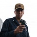 A U.S. Navy sailor wearing a USS Farragut DDG-99 cap and dark blue coveralls is holding a handheld device, likely a radio or measurement instrument. He appears focused and is examining the object in his hand. The background is overcast and white, suggesting an outdoor naval environment.