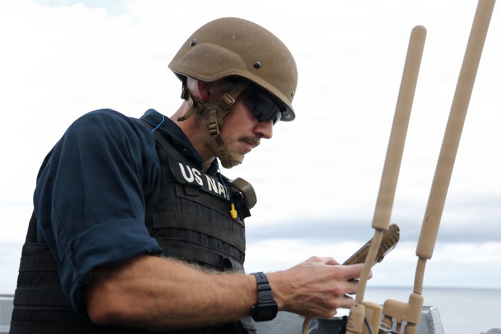 A US Navy personnel is pictured wearing a tactical vest labeled 'US NAVY', a tan/coyote brown ballistic helmet, and sunglasses. He appears to be operating or inspecting equipment with dual antenna-like protrusions, likely a radio or communications device. The setting appears to be aboard a naval vessel at sea, based on the visible horizon and water in the background.
