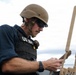 A US Navy personnel is pictured wearing a tactical vest labeled 'US NAVY', a tan/coyote brown ballistic helmet, and sunglasses. He appears to be operating or inspecting equipment with dual antenna-like protrusions, likely a radio or communications device. The setting appears to be aboard a naval vessel at sea, based on the visible horizon and water in the background.
