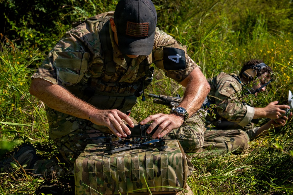 Two individuals in military-style multicam camouflage uniforms are positioned in a grassy outdoor area. The foreground figure is preparing or assembling a small drone/UAV on top of a camouflage-patterned case or bag. A second individual in the background appears to be holding a controller or device while wearing headgear.