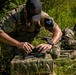 Two individuals in military-style multicam camouflage uniforms are positioned in a grassy outdoor area. The foreground figure is preparing or assembling a small drone/UAV on top of a camouflage-patterned case or bag. A second individual in the background appears to be holding a controller or device while wearing headgear.
