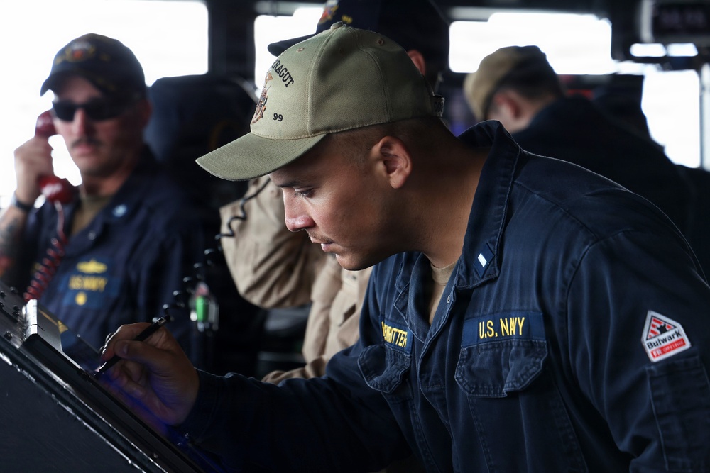 U.S. Navy personnel are shown working on the bridge of a naval vessel. The foreground sailor, wearing a 'Farragut 99' cap and U.S. Navy coveralls with a 'Bulwark' patch, is writing or recording data on a clipboard or console. Additional crew members are visible in the background, one of whom is speaking on a red telephone handset.