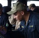 U.S. Navy personnel are shown working on the bridge of a naval vessel. The foreground sailor, wearing a 'Farragut 99' cap and U.S. Navy coveralls with a 'Bulwark' patch, is writing or recording data on a clipboard or console. Additional crew members are visible in the background, one of whom is speaking on a red telephone handset.