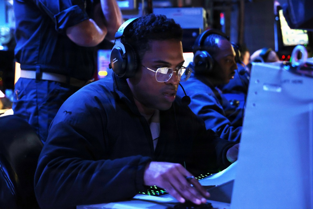 The image shows personnel working at computer workstations in a dimly lit, blue-toned operations or command center environment. The foreground subject is wearing headphones with a boom microphone and glasses, actively typing on a keyboard with illuminated green keys. The setting appears to be a military or naval combat information center (CIC) or similar tactical operations room.